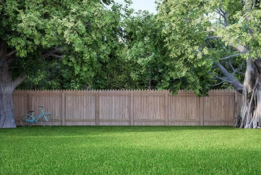 Wooden backyard fence enclosing a grassy yard, with large leafy trees overhead and a bicycle resting against the fence.