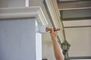 A person uses a paintbrush to apply white paint to crown molding on an exterior building column.