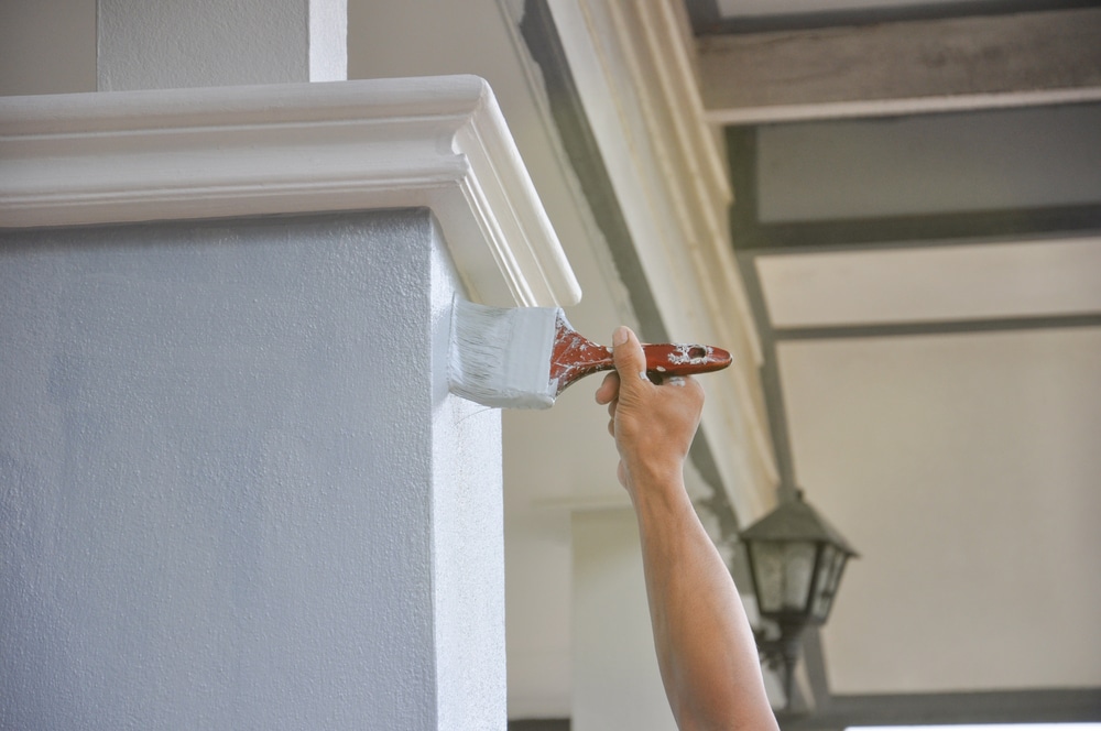 A person uses a paintbrush to apply white paint to crown molding on an exterior building column.