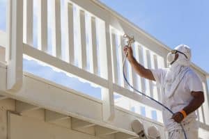 A house painter wearing protective clothing, a face mask, and goggles uses a spray gun to paint a white outdoor deck railing under a bright blue sky.